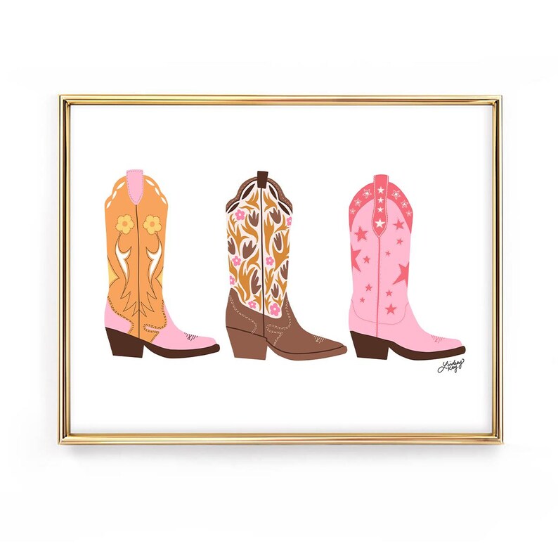 May include: A framed print featuring three colorful cowboy boots. The boots are in shades of orange, brown, and pink, each with unique floral or star patterns. The artwork is set against a white background.