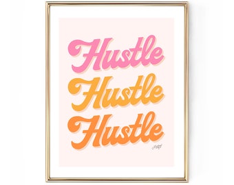 May include: A framed print with the word "Hustle" repeated three times in a retro font. The word is in shades of pink and orange on a light pink background, framed in gold.