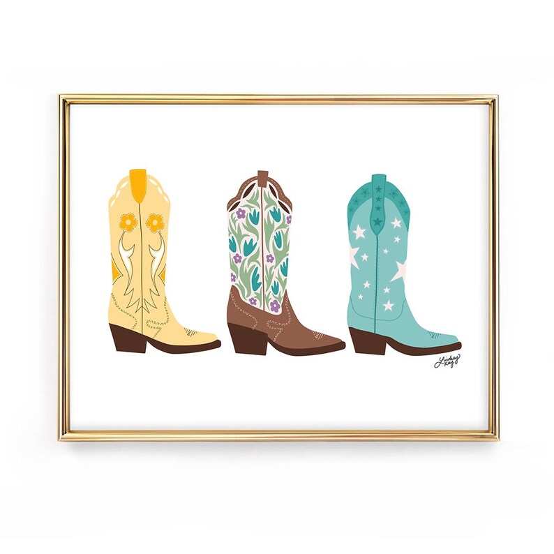 May include: A framed print featuring three colorful cowboy boots. The boots are yellow with orange flowers, brown with purple and green floral patterns, and turquoise with white stars. The artwork is set against a white background.