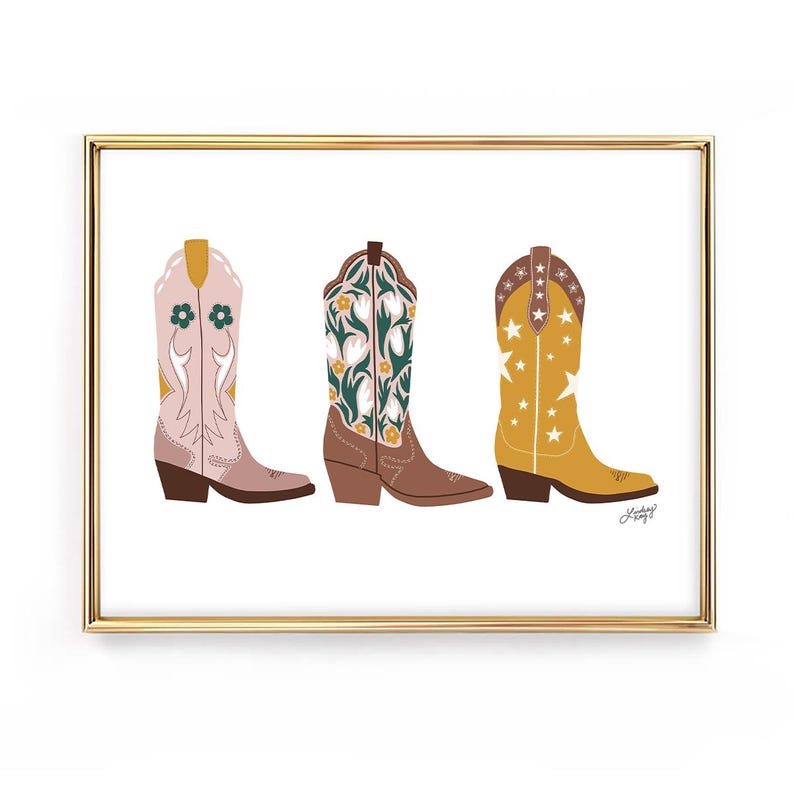 May include: A framed print featuring three colorful cowboy boots. The boots are in shades of pink, brown, and yellow, each with unique floral or star designs. The artwork is set against a white background.