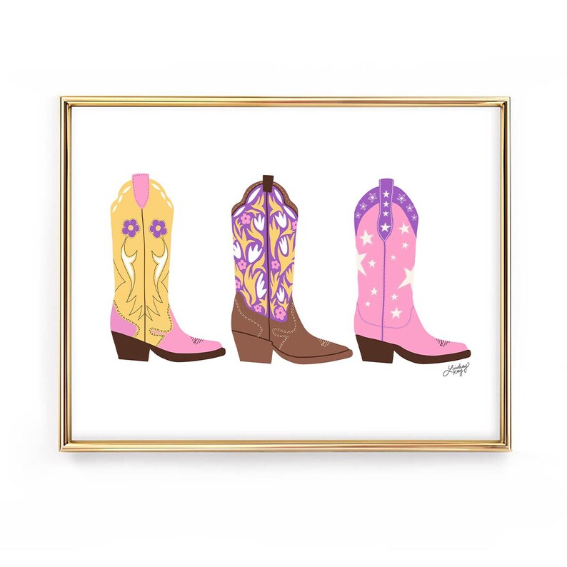 May include: A framed print featuring three colorful cowboy boots. The boots are in shades of yellow, brown, and pink, each with unique floral or star patterns. The artwork is set against a white background and framed in gold.