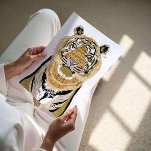 Puede incluir: Una impresi&oacute;n art&iacute;stica detallada de la cara de un tigre, con un collage de texto y varios tonos de marr&oacute;n, negro y blanco. Los ojos del tigre son azules. La obra de arte es sostenida por una persona vestida de blanco.