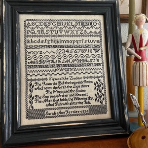 Puede incluir: Bordado de punto de cruz en blanco y negro enmarcado en un marco de madera negro. El bordado presenta el alfabeto, los números y un poema sobre los signos del zodíaco. El texto dice: "Signos del zodíaco. El carnero el toro los gemelos celestiales Y luego el cangrejo el león brilla La virgen y la balanza El escorpión el arquero y la cabra marina El hombre que sostiene la jarra de riego Y los peces con colas brillantes. Sarah Ann Fernley - 1826"