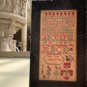 Könnte beinhalten: Ein gerahmter Kreuzstich-Sampler mit einem schwarzen Rahmen. Der Sampler zeigt ein farbenfrohes Alphabet, Zahlen und eine kleine Szene mit Vögeln und Blumen. Der Text auf dem Sampler lautet "Janet Grant AD/4/1866".