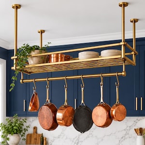 May include: A gold-toned metal ceiling-mounted pot rack featuring a top shelf holding a potted plant, copper bowls, and white plates. Six S-shaped hooks hang from the lower bar, supporting various copper and dark metal cookware pieces.
