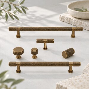 May include: A collection of antique brass cabinet hardware including two long bar pulls, one T-bar knob, two round knobs of different sizes, and one cylindrical knob. Each piece features a textured, knurled surface pattern throughout.