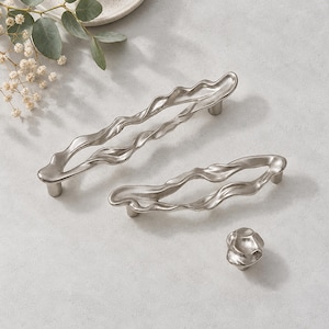 May include: Two silver-toned metal cabinet pulls with an elongated, wavy, organic shape and a matching small, round, textured cabinet knob. The hardware features a smooth, reflective finish with irregular, flowing contours.