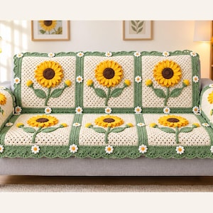 Puede incluir: Una funda de sofá de ganchillo color crema con grandes girasoles amarillos tridimensionales con centros marrones y tallos verdes. La funda está decorada con pequeños motivos de margaritas blancas en los bordes verdes.