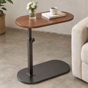 May include: An oval-shaped side table with a dark wood-grain top and a black metal base. The table features a single vertical support column with an adjustable height knob. A white mug and a book are placed on the tabletop surface.
