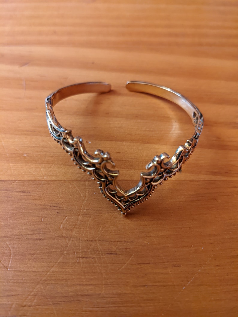 Pode incluir: Uma pulseira de punho de metal dourado com um design aberto. A pulseira apresenta um padr&atilde;o ornamental em estilo filigrana ao longo da banda e uma pe&ccedil;a central decorativa em forma de V. A pulseira &eacute; exibida em uma superf&iacute;cie de madeira.