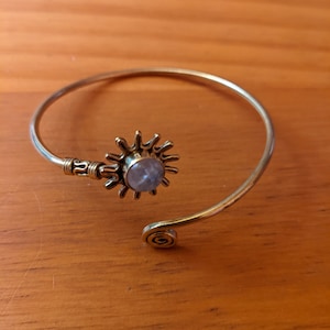 Op de afbeelding: Een goudkleurige armband met een zonmotief. De zon heeft een centrale, lichtgekleurde steen. De armband heeft een spiraalvormig ontwerp aan één uiteinde en een sierlijke sluiting aan de andere.