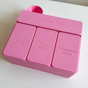 May include: A pink plastic organizer box with four distinct compartments. The top section is labeled Pregnancy Tests. The three lower sections are labeled Ovulation Strips, PdG Strips, and Pregnancy Strips. The box features a small circular cup-like attachment at the back.