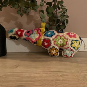 Può includere: Un elefante di peluche realizzato con motivi floreali all'uncinetto multicolori nelle tonalità del rosso, blu, giallo, verde e bianco. L'elefante presenta un nastro giallo brillante legato attorno al collo e una piccola coda bianca.