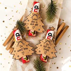 Könnte beinhalten: Drei Schokoriegel, verpackt in braunen Papierausstanzungen in Form von Weihnachtsbäumen. Jeder Anhänger zeigt eine handgezeichnete Schleife, Sterne, ein Herz und den deutschen Text: "Schön dass es dich gibt", "Du warst brav" und "Du bist ein Engel".