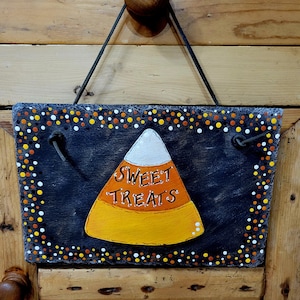May include: A black slate sign with a white polka dot border hangs from a brown knob. The sign features a hand-painted orange and yellow candy corn with the words "Sweet Treats" written in white.