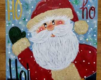 May include: Square wooden sign featuring a hand-painted Santa Claus with a white beard, red suit, and a green mitten. The words "HO ho Ho!" are painted in gold and black on a light blue background with white snowflakes.