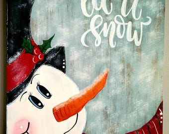May include: A hand-painted wooden sign with a snowman face and the text "Let it snow". The snowman has a black hat, a red scarf, and a carrot nose. The background is a light blue color with a white wash.