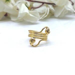 Puede incluir: Un anillo de metal en tono dorado con un diseño de espiral de varias capas. La banda consta de varios bucles de alambre paralelos que se envuelven alrededor del dedo, con cada extremo terminado en un pequeño adorno en forma de hoja.