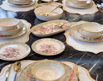 May include: A dinnerware set featuring white porcelain plates and bowls with a pink rose floral pattern, green decorative borders, and gold trim. The set includes matching placemats, gold-toned spoons, forks, and knives.