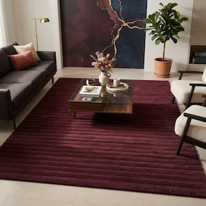 Puede incluir: Una alfombra rectangular grande de color burdeos intenso con una textura acanalada horizontal. La alfombra está situada en una sala de estar, centrada bajo una mesa de centro con tapa de cristal, un sofá gris oscuro y dos sillones color crema.