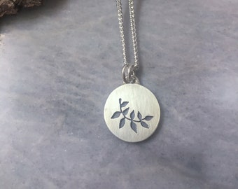 May include: A silver chain necklace with a round pendant featuring a cut-out design of a branch with leaves.