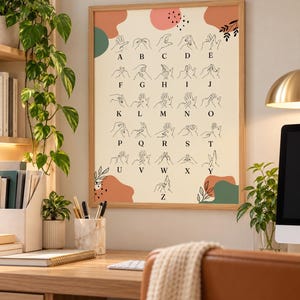 Può includere: Una stampa artistica rettangolare da parete in una cornice di legno chiaro con sfondo color crema e forme astratte color terracotta e verde. La stampa mostra l'alfabeto della lingua dei segni con disegni a linee nere di gesti e lettere dalla A alla Z.