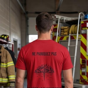 May include: A red short-sleeved t-shirt featuring black text that reads NE PANIQUEZ PAS at the top and JE GÈRE LE THERMOSTAT at the bottom, with a central graphic of flames above a horizontal line and a vertical needle pointing downward.