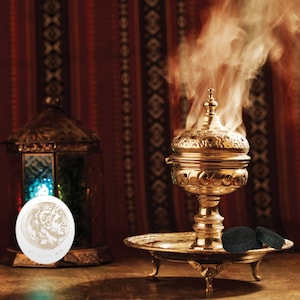 May include: A gold metal incense burner with ornate details sits on a silver tray with three legs. Smoke rises from the burner, creating a mystical atmosphere. Two black charcoal discs are on the tray.