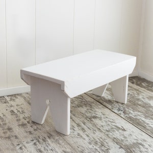 Puede incluir: Un taburete de madera blanco con un dise&ntilde;o sencillo. El taburete tiene una parte superior rectangular y cuatro patas. Las patas est&aacute;n ligeramente inclinadas y tienen un dise&ntilde;o de corte.