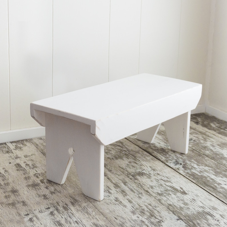 Puede incluir: Un taburete de madera blanco con un dise&ntilde;o sencillo. El taburete tiene una parte superior rectangular y cuatro patas. Las patas est&aacute;n ligeramente inclinadas y tienen un dise&ntilde;o de corte.