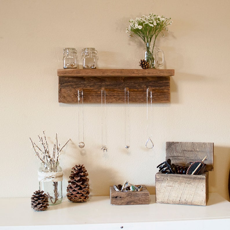 May include: Rustic wooden shelf with five hooks for hanging necklaces. The shelf has a distressed wood finish and is mounted on a white wall. There are two small glass jars on the shelf, and a vase with white flowers. There are also two pine cones and a small glass jar with twigs on the shelf.