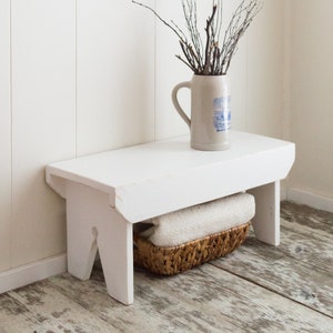 Puede incluir: Taburete de madera blanco con acabado desgastado. El taburete tiene un dise&ntilde;o sencillo con dos patas en la parte delantera y dos patas en la parte trasera. Una cesta tejida con una toalla blanca est&aacute; debajo del taburete.