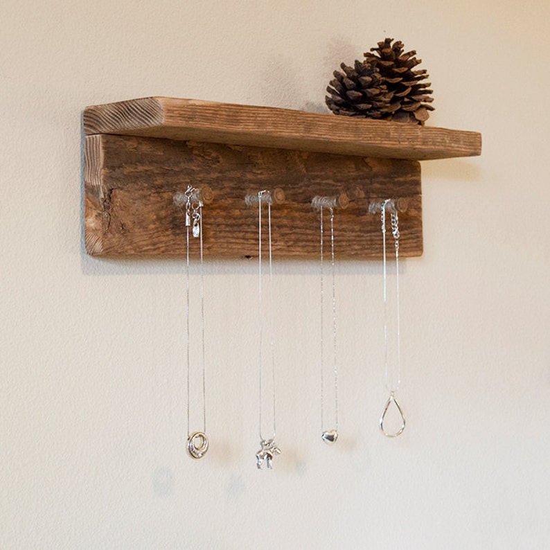 May include: Rustic wooden shelf with six hooks for hanging necklaces. The shelf is stained a dark brown and has a natural wood grain. There are five necklaces hanging from the hooks, each with a different pendant.