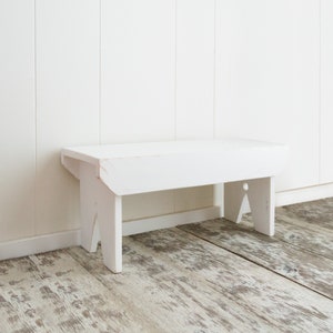 Puede incluir: Un banco de madera blanco con un dise&ntilde;o sencillo. El banco tiene una parte superior rectangular y dos patas a cada lado. El banco est&aacute; sentado en un suelo de madera clara.