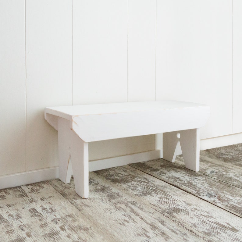 Puede incluir: Un banco de madera blanco con un dise&ntilde;o sencillo. El banco tiene una parte superior rectangular y dos patas a cada lado. El banco est&aacute; sentado en un suelo de madera clara.