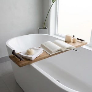 May include: A rectangular wooden bathtub tray with dark metal handles on each end. The tray holds a white folded washcloth with a round soap bar on top, an open book with white pages, and two cylindrical cream-colored candles on the right side.