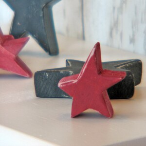 May include: Three wooden stars, two painted dark blue and one painted red, are arranged on a white surface. The red star is in the center and the blue stars are on either side.