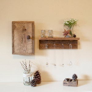 May include: Rustic wooden shelf with hooks for hanging necklaces, a small wooden cabinet with a knob, and two glass jars. The shelf is decorated with a small wooden star and a vase of white flowers.