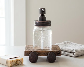 Peut inclure: Un distributeur de savon en verre transparent avec un dessus de pompe en bronze repose sur une petite plateforme en bois rustique avec des roues marron foncé. Une barre de savon et un gant de toilette gris sont également visibles.