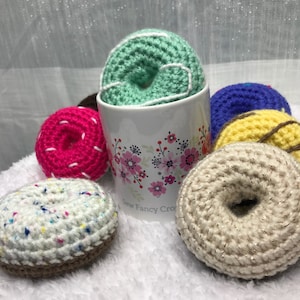 Puede incluir: Una colección de coloridos donuts de ganchillo en varios colores, incluyendo rosa, azul, amarillo y blanco, con chispas y glaseado decorativos. Un donut está dentro de una taza con estampado floral. El texto "Sew Fancy Crochet By Ashley Cosens" es visible.