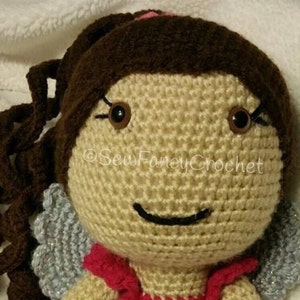 May include: A handmade crochet doll with brown hair styled in a ponytail and long, curly tendrils. The doll has brown eyes, a smiling face, and small silver wings. The doll is wearing a red top. The text "Sew Fancy Crochet" is visible.