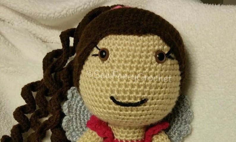 May include: A handmade crochet doll with brown hair styled in a ponytail and long, curly tendrils. The doll has brown eyes, a smiling face, and small silver wings. The doll is wearing a red top. The text "Sew Fancy Crochet" is visible.