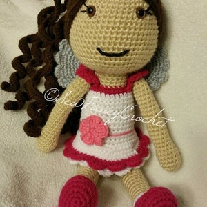 May include: A handmade crochet doll with brown curly hair, a beige face, and a white dress with red trim. The doll has pink shoes and a pink flower on the dress. The doll has gray wings and the text "@SewFancyCrochet".