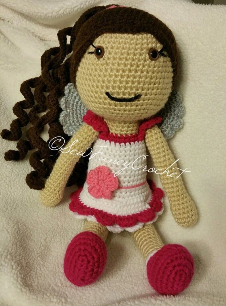 May include: A handmade crochet doll with brown curly hair, a beige face, and a white dress with red trim. The doll has pink shoes and a pink flower on the dress. The doll has gray wings and the text "@SewFancyCrochet".