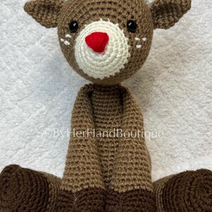 May include: A handmade crochet reindeer doll with a brown body, head, and legs. The reindeer has white antlers, a white muzzle, a red nose, and black eyes. The doll is sitting on a white surface.