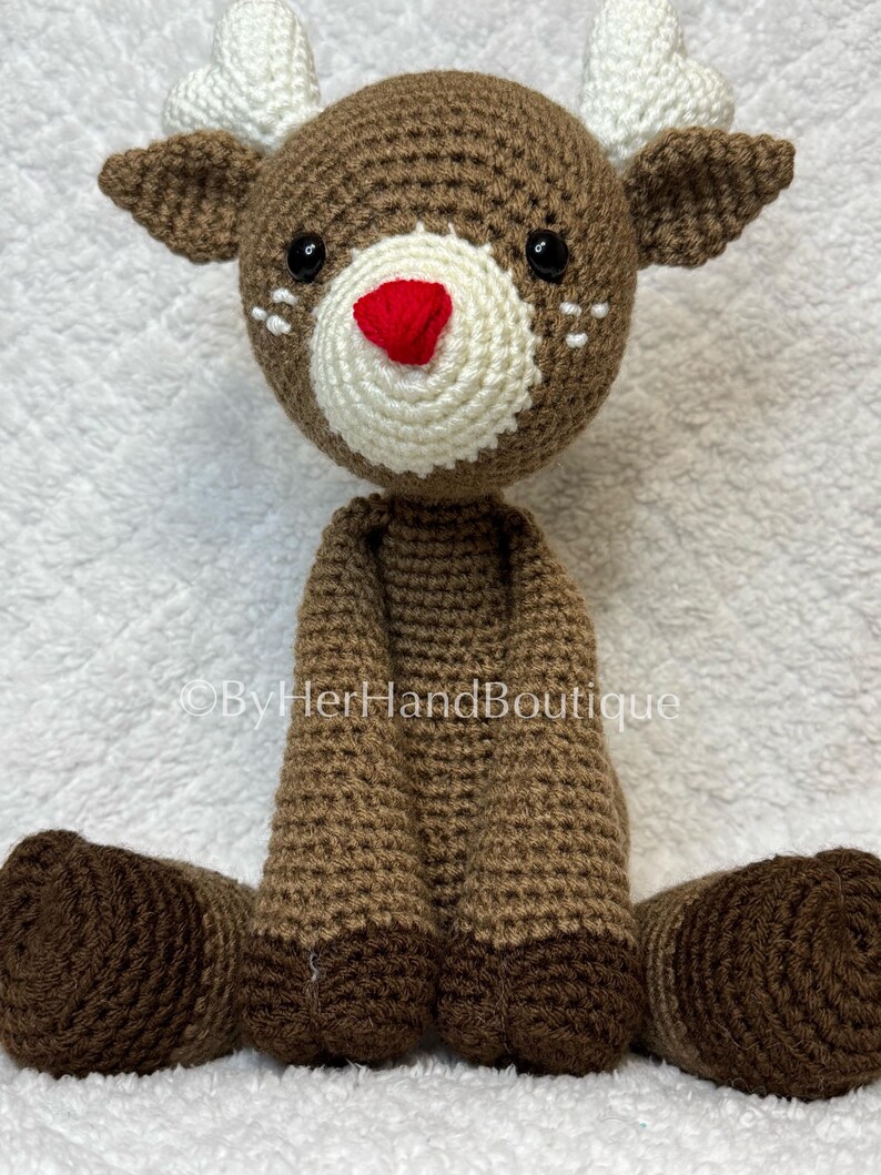 May include: A handmade crochet reindeer doll with a brown body, head, and legs. The reindeer has white antlers, a white muzzle, a red nose, and black eyes. The doll is sitting on a white surface.