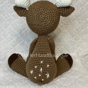 May include: A handmade brown crochet deer with white heart-shaped antlers. The deer has white star-shaped accents on its rear and brown hooves. The text "&copy;ByHerHandBoutique" is visible on the image.