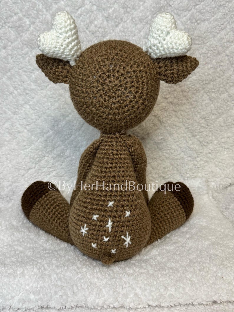May include: A handmade brown crochet deer with white heart-shaped antlers. The deer has white star-shaped accents on its rear and brown hooves. The text "&copy;ByHerHandBoutique" is visible on the image.