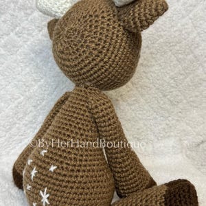 May include: A handmade brown crochet deer with white antlers and white star accents on its body. The deer has a rounded body, long legs, and a small tail. The deer is sitting on a white surface.