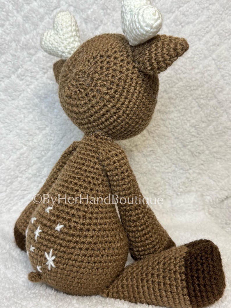 May include: A handmade brown crochet deer with white antlers and white star accents on its body. The deer has a rounded body, long legs, and a small tail. The deer is sitting on a white surface.